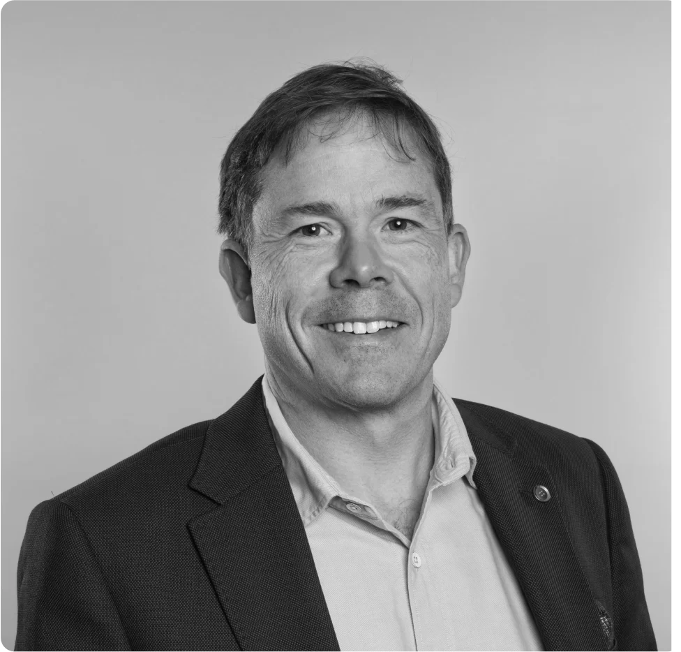A man in a suit jacket and collared shirt smiles at the camera against a plain background. The photo is in black and white.