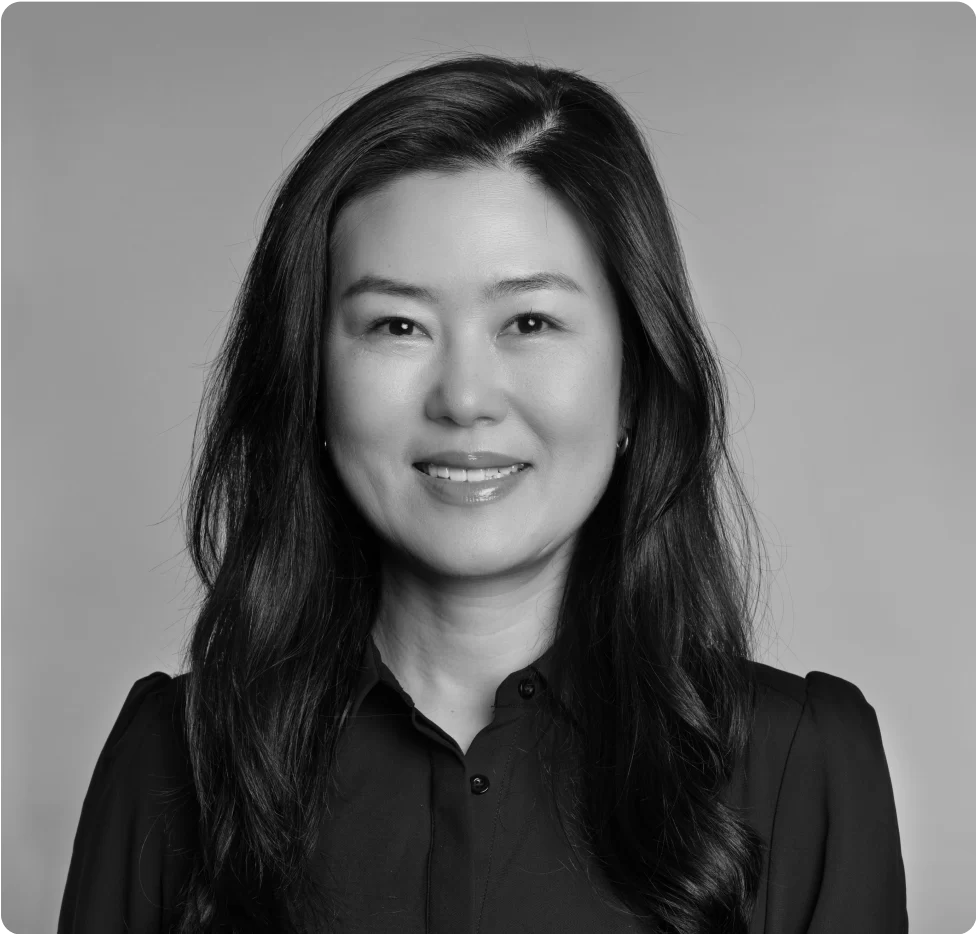A woman with long dark hair, wearing a dark button-up shirt, smiles at the camera against a plain background. The photo is in black and white.