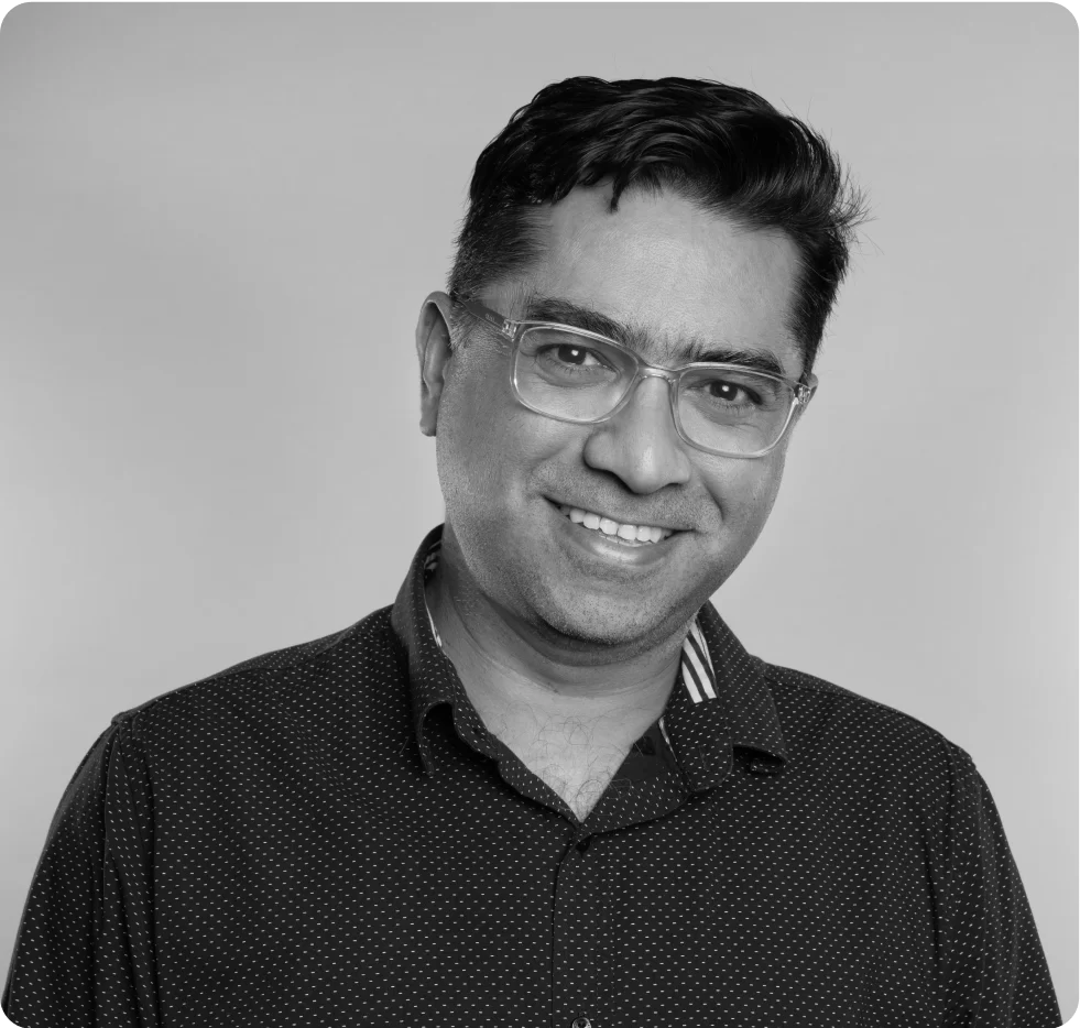 A man with short dark hair and glasses smiles at the camera. He is wearing a dark, patterned, button-up shirt. The background is plain and light-colored. The photo is in black and white.