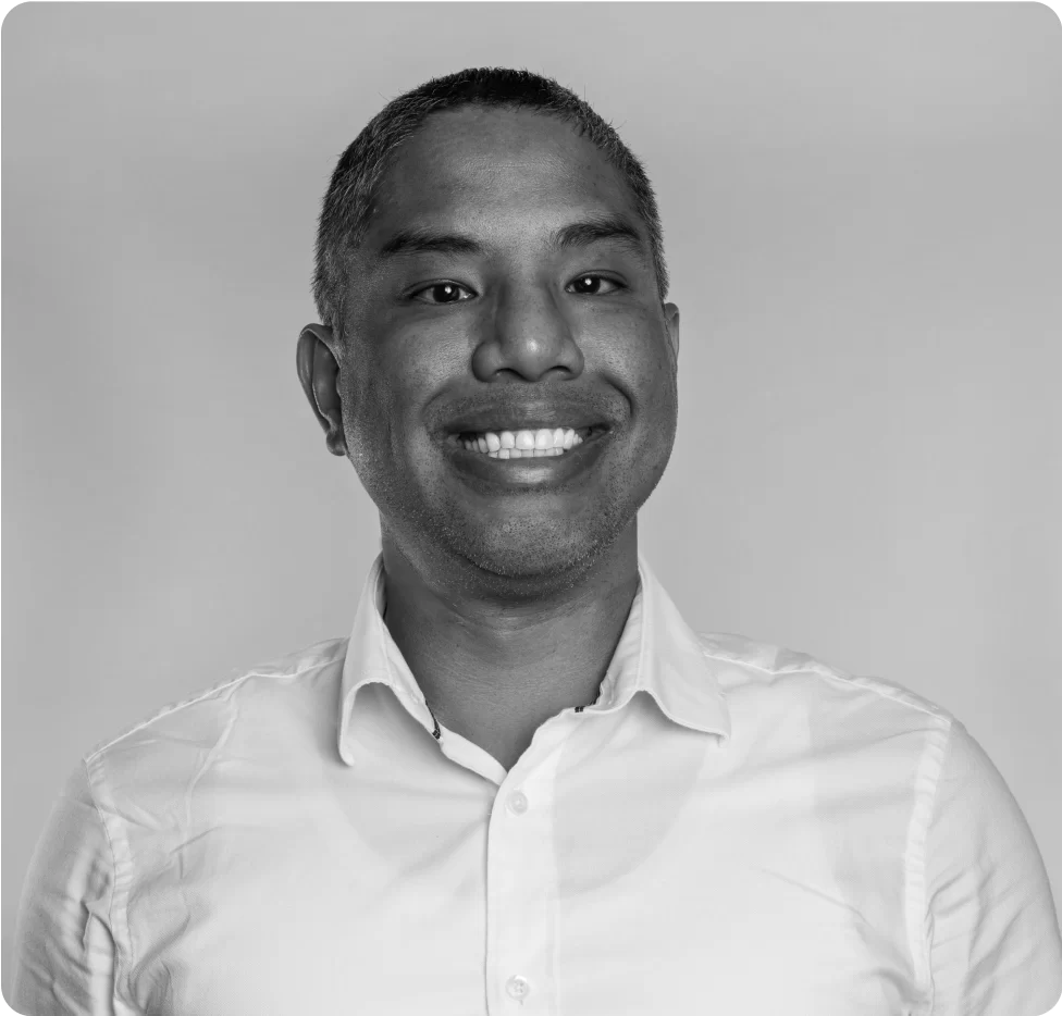 A man with short hair smiles, wearing a white collared shirt, against a plain light background. The photo is in black and white.