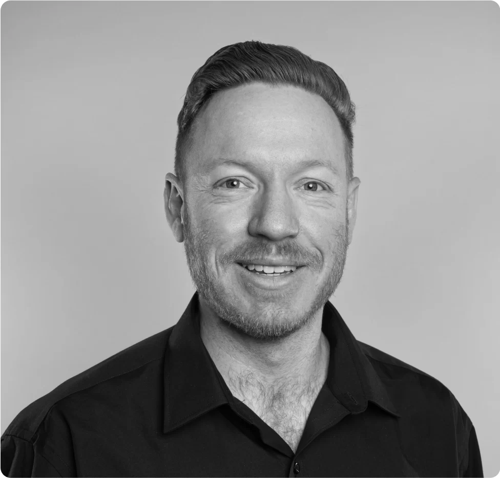 A man with short hair and a trimmed beard, wearing a dark collared shirt, smiles at the camera against a plain background.
