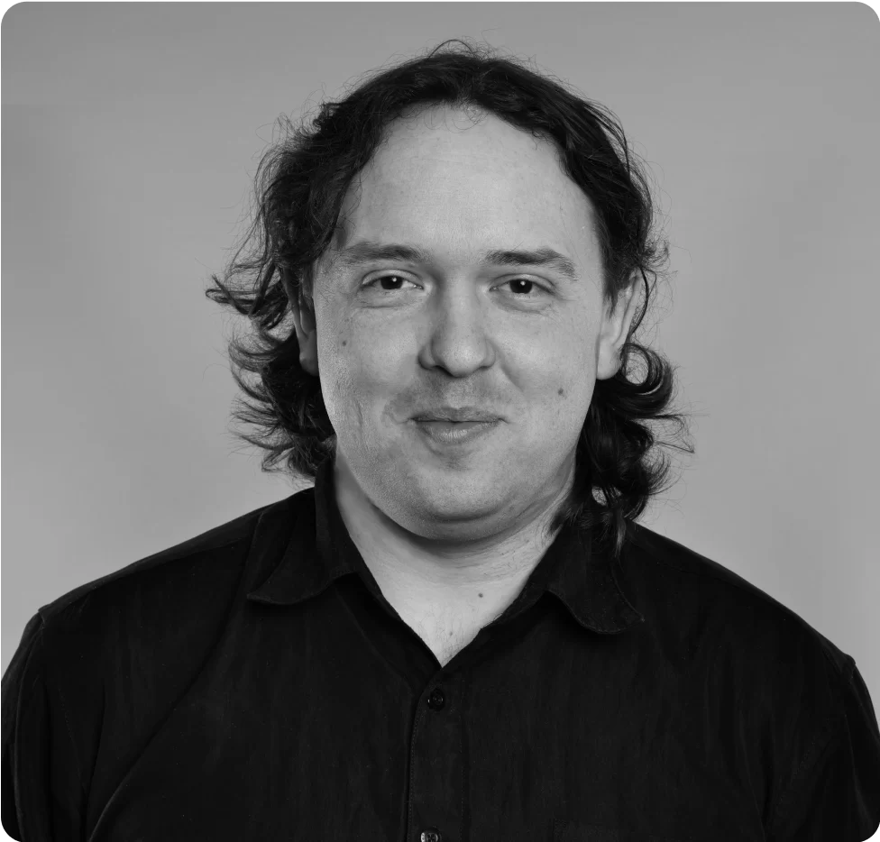 A man with shoulder-length dark hair wearing a dark button-up shirt faces the camera and smiles slightly, against a plain background.