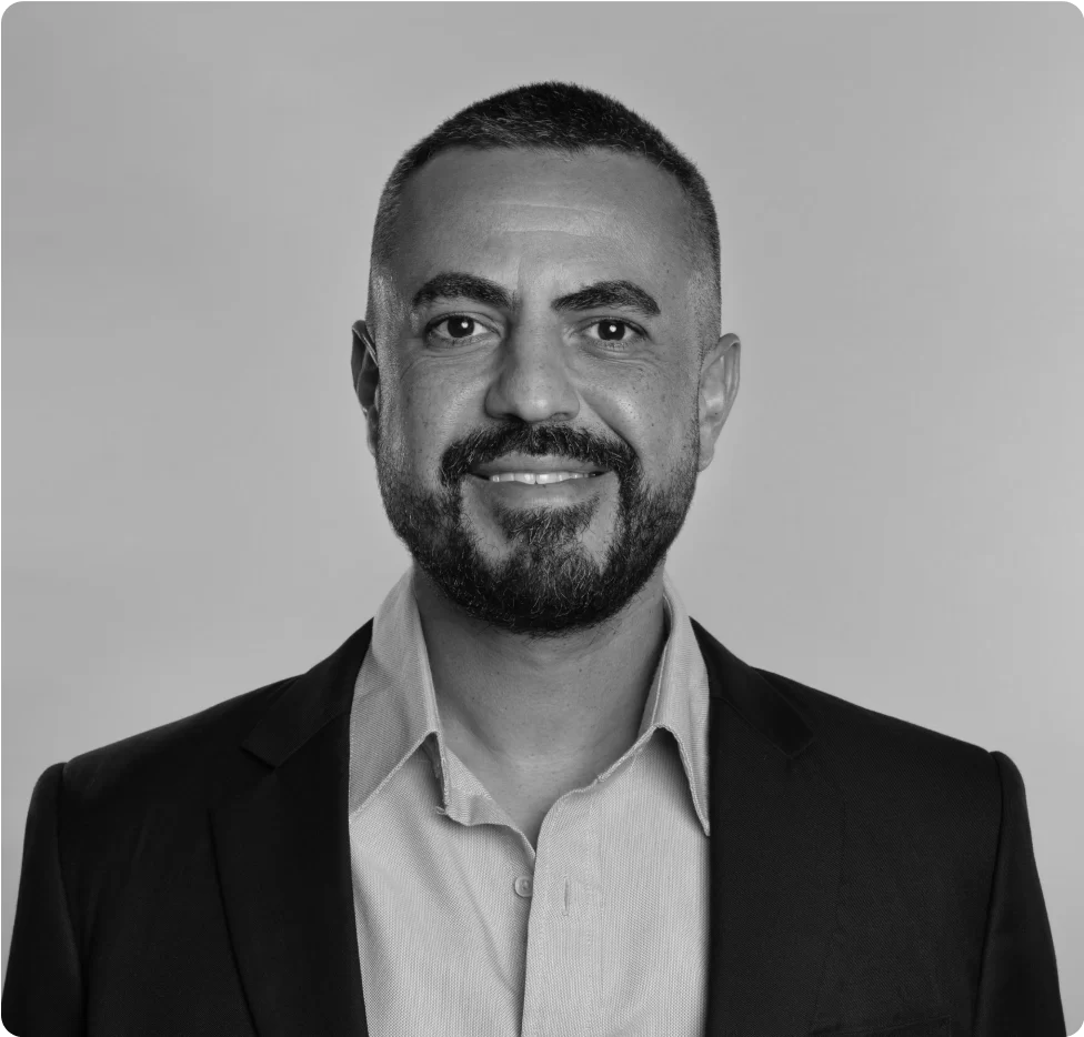 A man with short hair and a beard wearing a suit jacket and collared shirt, smiling at the camera against a plain background.