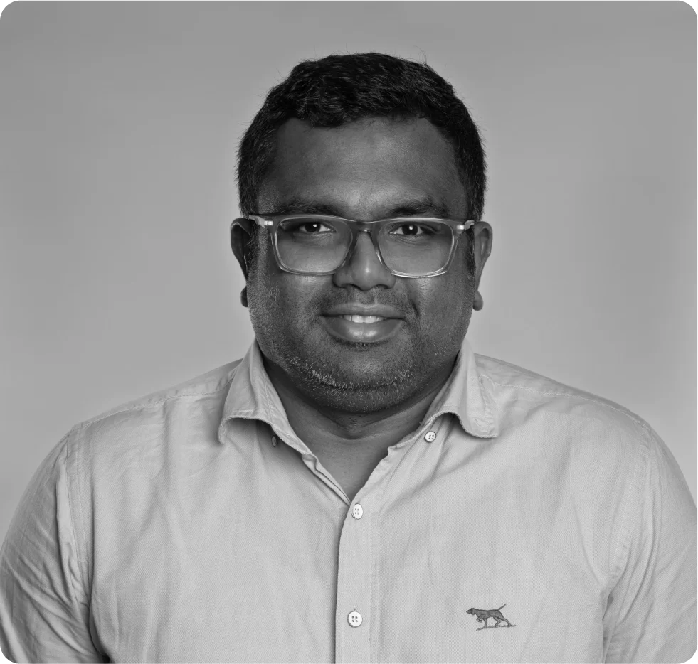 A man with short hair and glasses wearing a light-colored button-up shirt with an animal logo, smiling at the camera against a plain background.