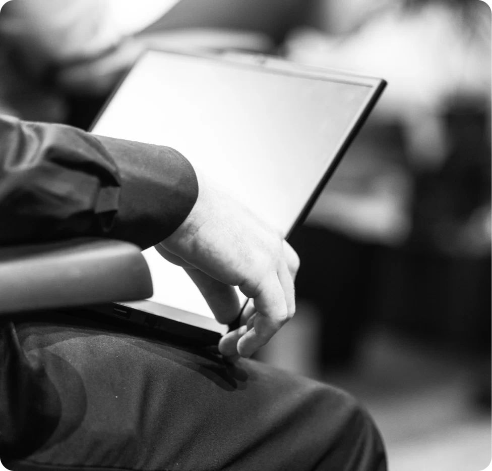 A person seated and holding an open laptop on their lap; the image is in black and white and focused on the hand and device.