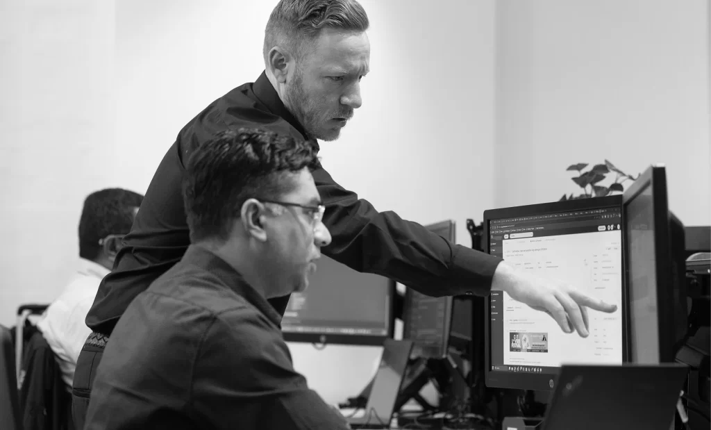 Two men in an office setting work at a computer; one is seated while the other stands next to him, pointing at the monitor and providing guidance.